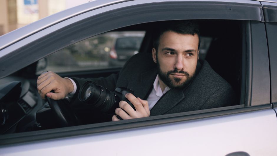 A bearded man in a car holding a camera, ready to take photos outdoors.